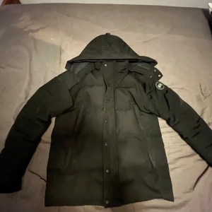 Svart dunjacka från Canada Goose - Säljer en svart dunjacka från Canada Goose med huva och knappar. Jackan har en broderad logotyp på ärmen och är perfekt för kalla dagar. Den är långärmad och har en stilren design.