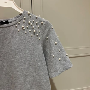 Grå t-shirt med pärldetaljer - Snygg grå t-shirt med korta ärmar och pärlor på axlarna. Som ny!
