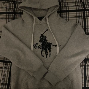 Ralph Lauren hoodie - Säljer en stilren grå hoodie från Polo Ralph Lauren med det ikoniska polospelartrycket på framsidan. Tröjan har en huva med vita snören och ribbade muddar vid ärmslut och nederkant. Den passar inte tyvärr inte mig då jag är åt det större hållet. Den är i storlek m men skulle säga att den krympt till s. Perfekt för en avslappnad stil. Skick 9/10