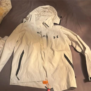 Vit vindjacka från Under Armour - Snygg vit vindjacka från Under Armour med huva och dragkedja. Jackan har svarta detaljer och praktiska fickor med dragkedjor. Perfekt för blåsiga dagar.