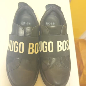 Sneakers  från Hugo Boss - Snygga svarta sneakers från Hugo Boss med bred resårrem över foten med logotyp. Skorna har en stilren design och är perfekta för en avslappnad look.
