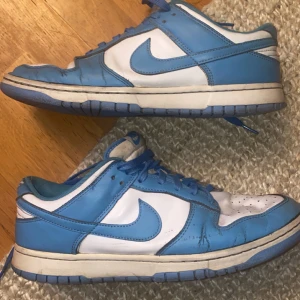 Nike Dunk i blått och vitt - Säljer ett par klassiska Nike Dunk sneakers i blått och vitt. Skorna har en låg profil med snörning och den ikoniska Nike-loggan på sidan. Perfekta för en avslappnad stil. Dom är dock lite creasade vilket man ser på bilderna