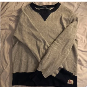 Grå tröja från Polo Ralph Lauren - Säljer en stilren grå tröja från Polo Ralph Lauren med mörkblå detaljer vid halsringningen och ärmsluten. Perfekt för en avslappnad look.