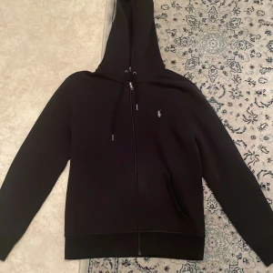 Svart hoodie från Ralph Lauren - Snygg svart hoodie från Ralph Lauren med dragkedja och huva. Den har en liten logga på bröstet och är perfekt för en avslappnad stil och vardags stil . Skriv om ni har frågor! Kan sänkas vid snabb affär!!