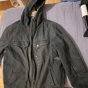 Svart jacka med huva - Snygg svart jacka med huva från Fifth Avenue. Jackan har dragkedja och flera fickor, inklusive en med knapp. Perfekt för en casual look. Den här slitage. Den kom så eftersom jag köpte det på second hand. 