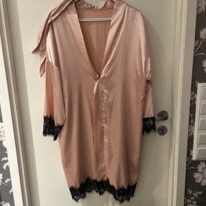 Babyrosa Rosa sidenklänning med spetsdetaljer - Elegant babyrosa klänning i siden med svarta spetsdetaljer vid ärmslut och nederkant. Morgonrock har en v-ringad design och trekvartsärmar, perfekt för en kväll