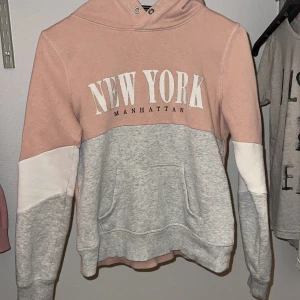 Rosa och grå hoodie med text - Säljer en snygg hoodie i rosa och grått med texten 'New York Manhattan' på bröstet. Tröjan har en stor ficka framtill och en bekväm huva.