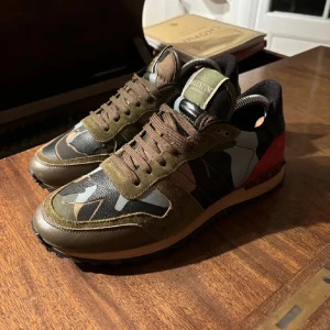 Valentino Garavani sneakers i camouflage - Snygga Valentino Garavani sneakers med ett coolt camouflagemönster i gröna och bruna toner. Skorna har röda detaljer på hälen och är tillverkade i en mix av skinn och mocka. De har snörning och en bekväm sula för extra komfort. Pris kan diskuteras!!