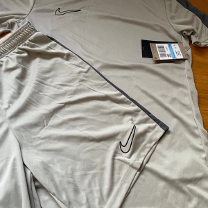 OANVÄND, med lapp kvar: Beige/grey sett från Nike - Snygga beige sett från Nike, shorts med elastisk midja och det klassiska Nike-logotypen i svart samt gråa detaljer på ärmarna och låren . Perfekta för träning eller en avslappnad dag. Materialet är lätt och andas bra. Buda gärna! 