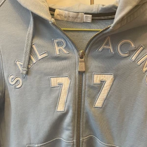 Ljusblå hoodie från Sail Racing - Säljer en ljusblå hoodie från Sail Racing med dragkedja och broderad text 'Sail Racing 77' på framsidan. Tröjan har en bekväm passform med långa ärmar och en praktisk huva med dragsko.