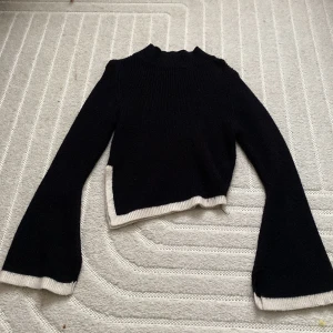 Svart stickad tröja med vita detaljer - Säljer en svart stickad tröja med vita kanter. Tröjan har långa ärmar och en croppad design som ger en modern look. Perfekt för kyligare dagar.