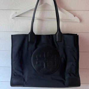 Svart axelväska från Tory Burch - Snygg svart väska från Tory Burch! Modell: Ella Tote. Höjd: 34cm, Längd: 43 cm, Bredd: 15 cm. Väskan är använd men i superfint skick, lite skav på botten (se bild) men annars inget att anmärka🙌
