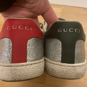 Glittriga sneakers från Gucci - Snygga glittriga sneakers från Gucci med kardborreband. Skorna har en silverglittrig yta med röd och grön detalj på hälen. Perfekta för att ge din outfit en extra touch av glamour. Finns kartong till, självklart äkta, inköpta på Harrods i London.
