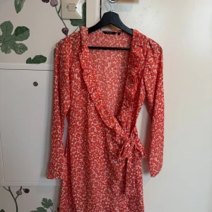 Röd blommig omlottklänning från Ally Bell - Charmig röd omlottklänning med blommigt mönster från Ally Bell. Klänningen har långa ärmar och en volangdetalj längs halsringningen. Perfekt för en somrig look!