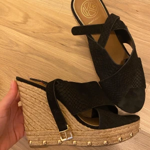 Svarta kilklackar från Kurt Geiger - Snygga svarta kilklackar från Kurt Geiger med flätad sula och gulddetaljer. Skorna har en öppen tå och ett justerbart spänne vid ankeln. Perfekta för sommarens alla tillfällen.