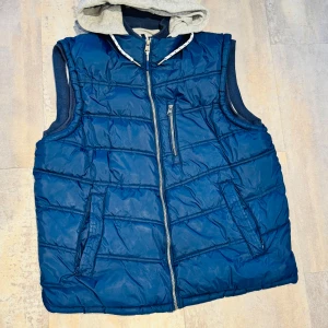 Blå quiltad dunväst med grå huva - Snygg blå quiltad dunväst med grå huva och dragkedja framtill. Västen har praktiska fickor och en sportig look, perfekt för kyliga dagar. Den grå insidan ger en skön kontrast och extra värme.