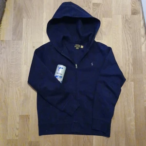 Mörkblå hoodie från Ralph Lauren - Säljer en snygg mörkblå hoodie från Ralph Lauren med dragkedja och huva. Den har det klassiska Ralph Lauren-logotypen broderad på bröstet. Perfekt för en avslappnad stil. Säljer eftersom den alltid varit för liten. Använd 5-10 gånger.