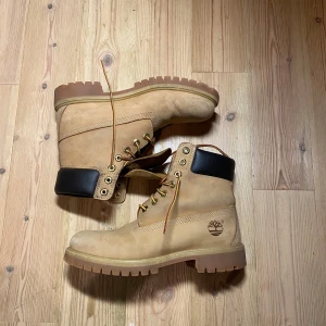 Beige boots från Timberland - Säljer ett par klassiska beige boots från Timberland med svart vadderad krage och robust snörning. Perfekta för höst och vinter med slitstark sula och ikonisk logga på sidan. Skicket är 8/10 allt det behövs är en tvätt så ser dem fräsha ut