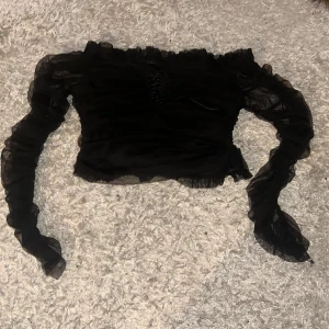 Svart offshoulder topp från Trendyol Trendyol - Snygg svart offshoulder topp från Trendyol med volangdetaljer och transparenta långa ärmar. Perfekt för en stilfull look med en touch av elegans. Passar utmärkt till både jeans och kjol.