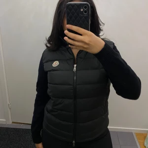Svart cardiganen från Moncler - Snygg svart dunväst från Moncler med dragkedja och logotyp på bröstet. Storlek L men liten i storleken. Perfekt för kyligare dagar och enkel att matcha med olika outfits. Prislapp följs med