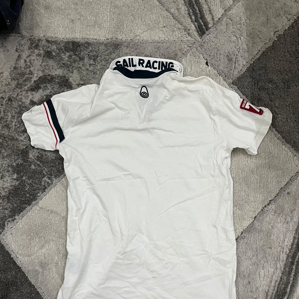 Snygg vit t shirt från Sail Racing med korta ärmar och stilrena detaljer i marinblått och rött. Tröjan har en klassisk krage och logotyp på bröstet samt text på kragen. Perfekt för en avslappnad och sportig look.. T-paidat.