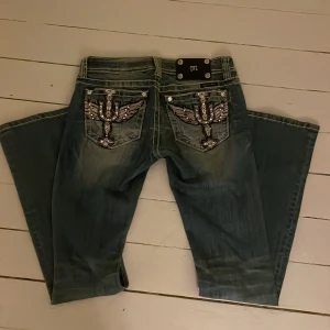 Miss me jeans - Snygga blå jeans från Miss Me med unika broderade detaljer på bakfickorna. Jeansen har en låg midja och en bootcut-stil. Innerben: 75cm och tvärs över är midjemåttet 35cm.