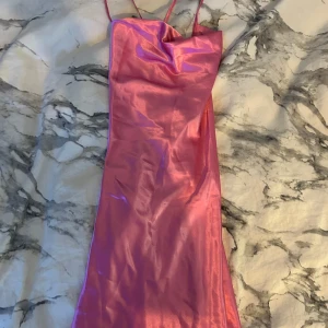 Rosa klänning - Snygg rosa klänning med smala axelband och en elegant drapering framtill. Perfekt för en somrig look eller en utekväll. Klänningen har en glansig finish som ger en lyxig känsla.