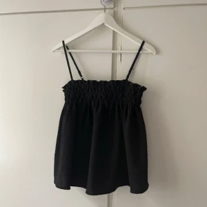Svart topp - Snygg svart axelbandslös topp med smockdetaljer upptill och justerbara axelband. Perfekt för en stilren look.
