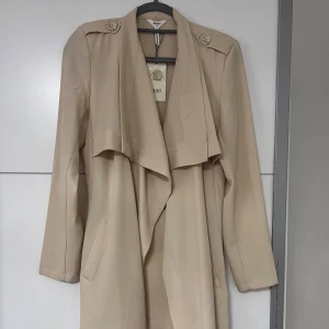 Beige trenchcoat med bälte - Snygg beige trenchcoat med klassisk design och bälte i midjan. Jackan har långa ärmar och dekorativa knappar på axlarna. Perfekt för en stilren look. (Kappan är ljusare än vad den ser ut på bild 1 och 2) så kika på tre sista bilderna. 