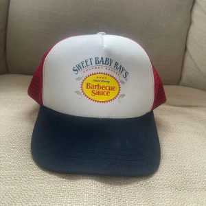 Sweet Baby Ray's keps - Cool keps med Sweet Baby Ray's logga. Den har en vit framsida med röd och gul logga, röd mesh på sidorna och en blå skärm. 