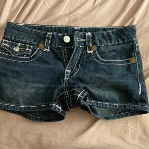 Mörkblå jeansshorts från True Religion - Snygga mörkblå jeansshorts från True Religion med kontrasterande ljusrosa sömmar och klassiska femficksdesign. Shortsen har dekorativa bakfickor med knappar och en låg midja för en avslappnad look. Knappt använda. Säljer pga för små 