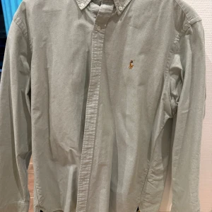 Grön skjorta från Ralph Lauren - Snygg grön skjorta från Ralph Lauren med klassisk krage och broderad logga på bröstet. Skjortan har långa ärmar och knappar framtill. Perfekt för en stilren look.