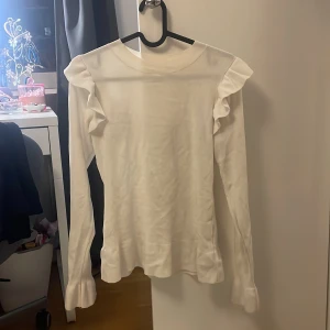 Vit långärmad topp med volangdetaljer - Säljer en stilren vit långärmad topp med volangdetaljer på axlarna. Perfekt för en elegant look. Toppen har en rund halsringning och är gjord i ett mjukt material.
