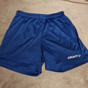 Blå shorts från Craft - Säljer ett par blå shorts från Craft i storlek S. De har en elastisk midja med snörning för justerbar passform. Perfekta för träning eller andra aktiviteter.