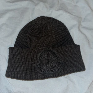 Svart stickad mössa från Moncler - Snygg svart stickad mössa från Moncler med broderad logga framtill. Mössan har en klassisk design med en uppvikt kant och en randig detalj i rött, vitt och blått på baksidan. Perfekt för att hålla värmen under kyliga dagar. Som ny pris kan diskuteras isåfall kom dm 