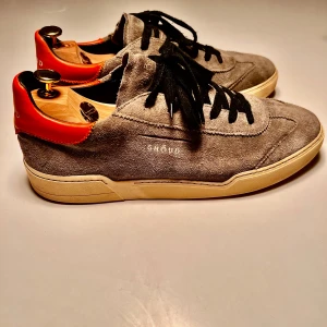 Grå sneakers från Ghoud (Ghōud) - Snygga grå sneakers i mocka från Ghoud med orange detaljer på hälen. Skorna har svarta snören och en vit sula som ger en stilren look. Perfekta för en casual stil.