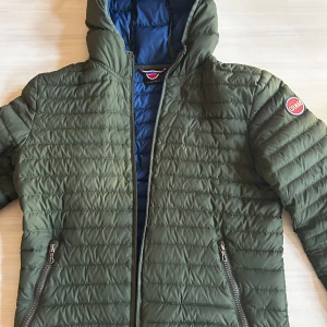 Grön quiltad jacka från Colmar - Snygg grön quiltad jacka från Colmar med blått foder. Jackan har en dragkedja framtill och en huva för extra skydd. Perfekt för kyligare dagar. Tyvärr är dragkedjan trasig men kan enkelt åtgärdas hos skräddare 🙌💯