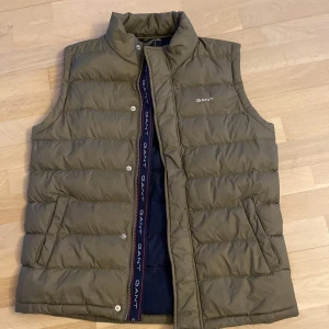 Olivgrön dunväst från GANT - Säljer en snygg olivgrön dunväst från GANT. Västen har en hög krage och stängs med dragkedja och knappar. Perfekt för lager-på-lager under kyliga dagar. Passar ungdomar i åldern 13-14 år.