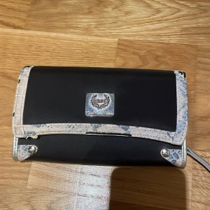 Svart plånbok med detaljer  - Säljer en plånbok i svart med silverdetaljer och ormmönstrad kant. Den har flera fack för kort och en dragkedja för mynt. Plånboken har flera små skador vilket man kan se på bilderna.