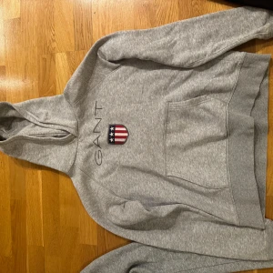 Grå hoodie från GANT - Säljer en stilren grå hoodie från GANT med en klassisk logga och ett emblem på bröstet. Tröjan har en stor ficka framtill och en bekväm huva. Perfekt för en avslappnad stil. Sparsamt använd, är i barnstorlek 170, 15år