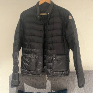 Svart pufferjacka från Moncler - Säljer en stilren svart pufferjacka från Moncler. Jackan har en quiltad design med dragkedja och knappar framtill. Perfekt för kyligare dagar. Finns en liten defekt bredvid fickan som är övertäckt med en lapp(väldigt liten), men annars är jackan i fint skick. Storlek 1/s men passar xs