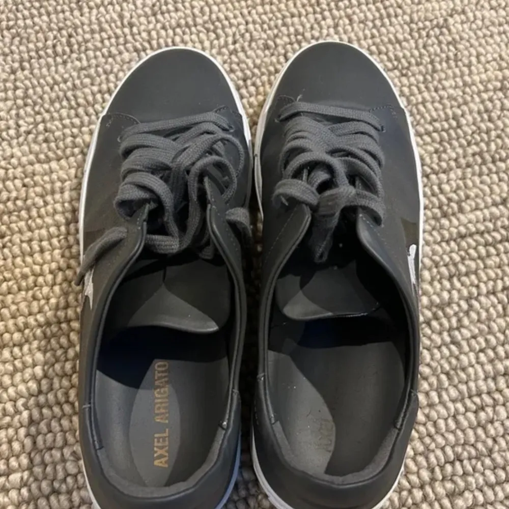 Snygga grå sneakers från Axel Arigato med vit sula och svart detalj på sidan. Skorna har en stilren design med snörning och en vit fågelbroderi som ger en unik touch. Pris kan diskuteras vid snabb affär!. Kengät.