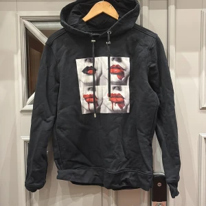 Limitato hoodie - Snygg svart hoodie med ett unikt tryck av röda läppar på framsidan. Tröjan har en justerbar huva med snören och långa ärmar. Perfekt för en cool och avslappnad stil. Märket är Limitato o nypriset ligger på 2500kr