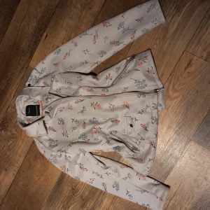 Blommig jacka från Yessica - Snygg blommig jacka från Yessica i ljusgrått med färgglada blommor i rosa och blått. Jackan har en dragkedja framtill och en klassisk krage. Perfekt för vår och sommar! Sparsamt använd. 