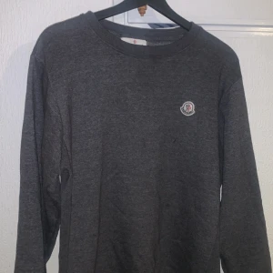 Grå tröja från Moncler - Säljer en stilren grå tröja från Moncler med rund hals och långärmad design. Tröjan har en liten logotyp på bröstet och är perfekt för en avslappnad look. Passar bra till både jeans och chinos.