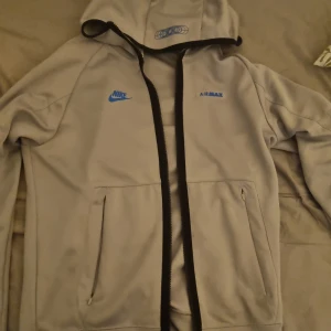 Nike hoodie - Säljer en grå hoodie från Nike med blå Air Max-logga. Den har dragkedja framtill och två fickor. Perfekt för en avslappnad stil.