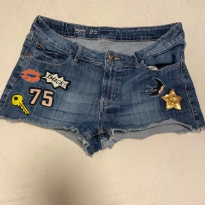 Jeansshorts med patches från Lindex - Coola jeansshorts från Lindex med olika patches som stjärna, fågel och text som 'SMACK' och 'OMG'. De har en fransig kant och är perfekt för en avslappnad stil. Märket Detroit Denim syns på knappen.