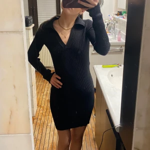 Svart ribbad klänning - Snygg svart ribbad klänning med lång ärm och v-ringad krage. Perfekt för en stilren look. Klänningen har en tight passform som framhäver figuren.