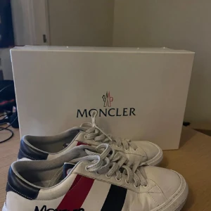 Vita sneakers från Moncler - Säljer ett par vita sneakers från Moncler med snörning och ränder i rött och blått på båda sidor. Har använts mycket men är fortfarande i användbart skick. Notera att sulorna under skorna är slitna (sista bilden).