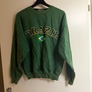 Grön sweatshirt från Nicks World - Säljer en grön sweatshirt från Nicks World med texten 'Brazil' och en brasiliansk flagga på framsidan. Tröjan har långa ärmar och ribbade muddar vid ärmslut och nederkant. Perfekt för en avslappnad stil!